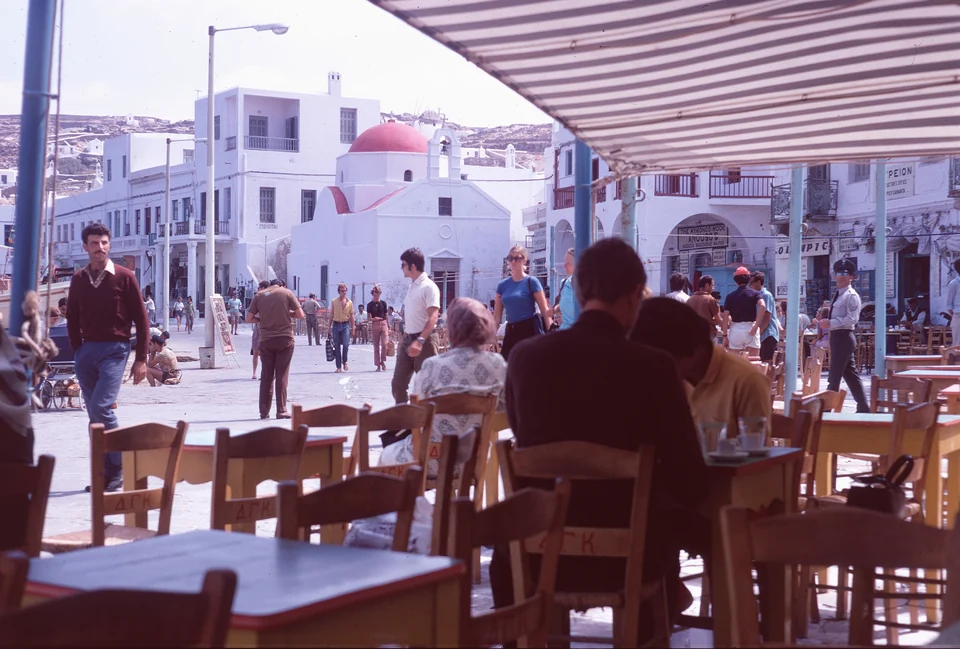 People seated at café tables under a striped awning beside a busy waterfront