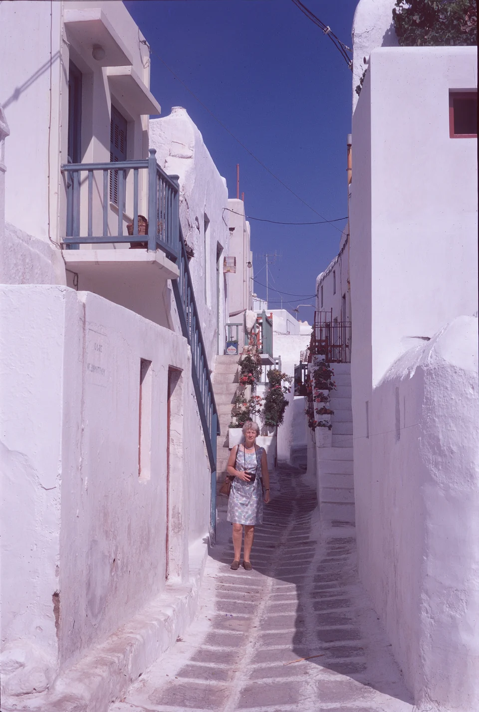 A person in a narrow lane between white buildings, balconies and exterior stairs