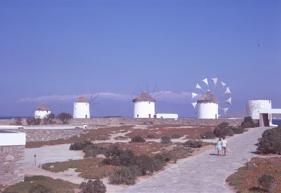 White windmills above stone walls and a path, with sea on the horizon