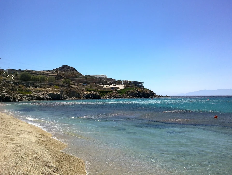 Άμμος και γαλαζοπράσινα νερά κάτω από βραχώδες ακρωτήρι με κτίρια στην παραλία Paradise