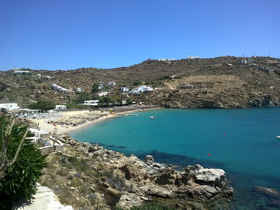Η παραλία Super Paradise από την απέναντι πλευρά του όρμου, με άμμο, ξαπλώστρες και τον βραχώδη λόφο γύρω της